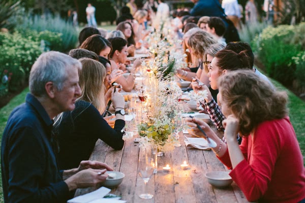 Guests of every age eating and talking along a long wooden table in a garden at dusk.