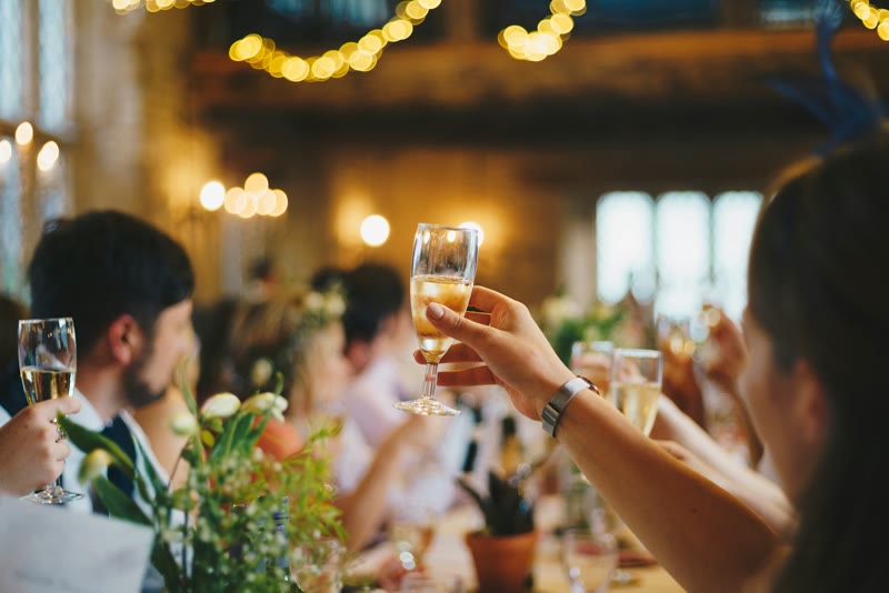 Wedding guests raising their glasses for a toast at a long table under warm string lights.
